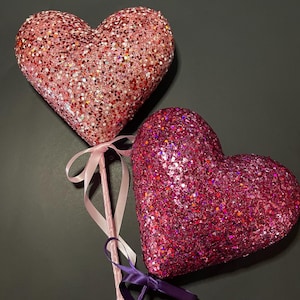 May include: Two glittery heart-shaped decorations on sticks with pink and purple ribbons. The hearts are pink and red.