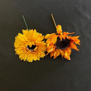 May include: Two artificial sunflowers with yellow petals and brown centers. The sunflower on the left has a lighter yellow color and a more defined center. The sunflower on the right has a darker yellow color and a more rounded center.