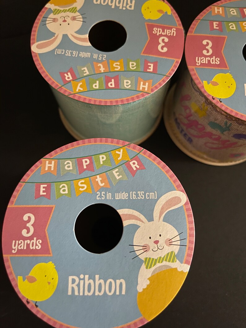 K&ouml;nnte beinhalten: Drei Spulen blaues und wei&szlig;es Band mit einem Happy Easter-Motiv. Jede Spule zeigt ein Kaninchen, ein gelbes K&uuml;ken und den Text "Happy Easter" in bunten Bannern. Das Band ist 2,5 Zoll breit und 3 Yards lang.