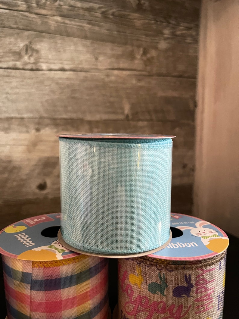 K&ouml;nnte beinhalten: Drei Rollen Band mit Ostern-Motiven. Die obere Rolle ist hellblau, die mittlere Rolle hat ein kariertes Muster in Pink, Blau und Gelb, und die untere Rolle hat einen braunen Jute-Hintergrund mit einem wei&szlig;en Hasen und dem aufgedruckten Text "Frohe Ostern".