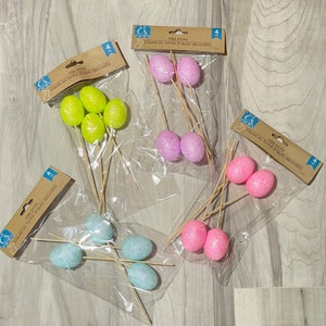 May include: Four packages of Easter egg picks. Each package contains four foam eggs on sticks. The eggs are in different colors: pink, light blue, green, and purple.