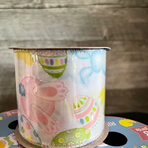 K&ouml;nnte beinhalten: Eine Rolle wei&szlig;es Satinband mit glitzernder Oberfl&auml;che. Das Band zeigt ein sich wiederholendes Muster aus rosa Hasen, pastellfarbenen Eiern und blauen V&ouml;geln. Das Band ist 2,5 Zoll breit und befindet sich auf einer Papprolle.