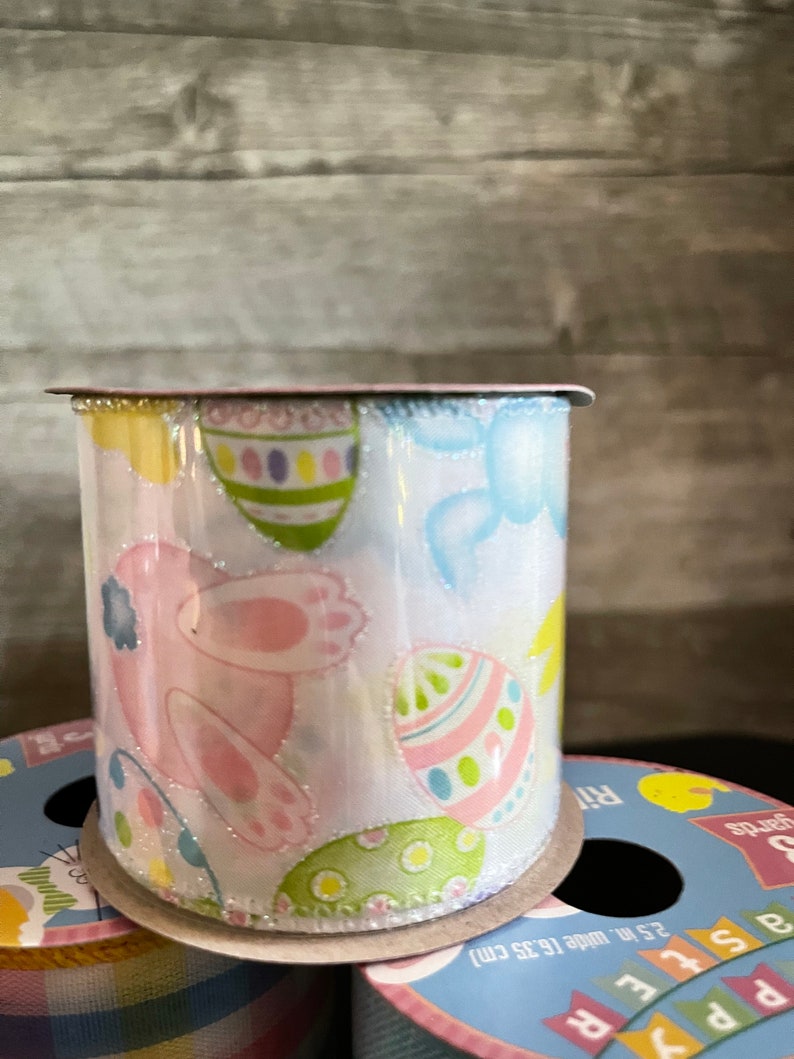 K&ouml;nnte beinhalten: Eine Rolle wei&szlig;es Satinband mit glitzernder Oberfl&auml;che. Das Band zeigt ein sich wiederholendes Muster aus rosa Hasen, pastellfarbenen Eiern und blauen V&ouml;geln. Das Band ist 2,5 Zoll breit und befindet sich auf einer Papprolle.