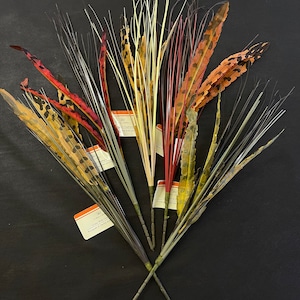 May include: A bouquet of faux grass stems with brown, black, red, and white feathers. The stems are tied together with twine and have small tags attached to them. The tags have text that reads "Grass with Feathers Bouquet of 6 stems".