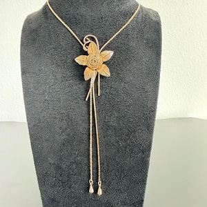 Puede incluir: Un collar tipo lazo en tono dorado con un colgante central de flor con pétalos y hojas texturizados. El collar tiene una cadena larga y delgada que se extiende hacia abajo desde la pieza central floral, terminando en dos pequeños dijes dorados.