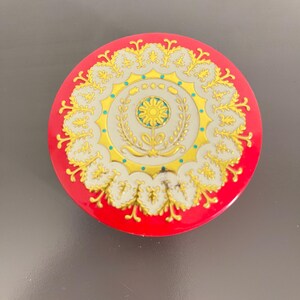 May include: A round, red decorative tin with a gold and white floral design. The center features a yellow flower within a wreath. The tin has a glossy finish and is set against a gray background.