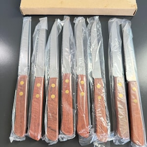 May include: A set of eight steak knives with serrated silver-tone metal blades and brown wooden handles secured by two brass rivets. Each knife is individually wrapped in clear plastic, and the set is accompanied by a rectangular cardboard box.