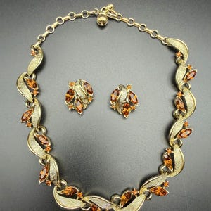 Pode incluir: Um conjunto de colar e brincos de pressão em metal dourado, com elos texturizados em forma de folha e strass cor de âmbar em lapidação marquise. O colar inclui uma corrente ajustável com um fecho redondo, combinando com os brincos.