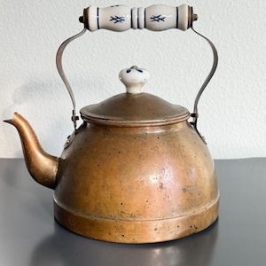 May include: A copper tea kettle with a rounded body and a curved spout. The lid features a white ceramic knob with blue floral accents. The handle is made of white ceramic with blue cross-hatch patterns, supported by two curved metal brackets.