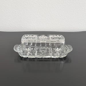 May include: A clear glass butter dish with a lid, featuring a rectangular shape and a textured, grid-like pattern. The dish has small handles and the lid has a decorative top. The butter dish is displayed on a dark surface.