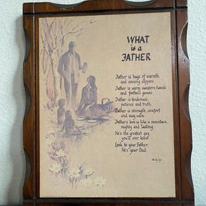 May include: Wooden framed print with the text "WHAT is a FATHER" and a poem about fathers. The artwork depicts a family scene in a park. The frame has a dark brown finish and a metal loop for hanging.