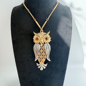 May include: A gold and silver-toned owl pendant necklace featuring a textured body with a diamond-patterned chest, smooth metallic wings, and two small amber-colored circular stones for eyes, suspended from a matching gold-toned metal chain.