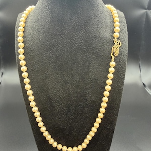 May include: A strand of cream-colored pearls displayed on a black mannequin. The necklace is fastened with a gold-colored clasp. The pearls are evenly sized and have a subtle luster. The necklace is a classic piece of jewelry.