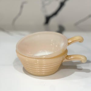 Puede incluir: Dos cuencos apilados, de color melocotón, de estilo vintage, con asas. Los cuencos tienen un diseño acanalado en el exterior y un interior liso. Las asas son curvas y del mismo color que los cuencos. Los cuencos están sobre una superficie blanca.