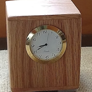 May include: A wooden clock with a gold-rimmed face and black hands. The clock is made of light brown wood and has a square shape. The clock face has black numbers and the words "Quartz" printed on it.