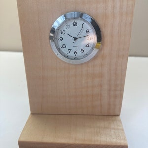 May include: A small, round, silver clock with black numbers and hands, mounted on a wooden stand. The clock face reads "QUARTZ".