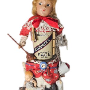 May include: A whimsical folk art doll made from a McCormick's Rubbed Sage can, featuring a doll head, red and white clothing, and miniature accessories. The doll has a metal funnel hat and a small doll at the base.