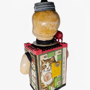 May include: A mixed-media sculpture featuring a vintage doll head topped with a dark wooden knob and a metal lid. The rectangular body is covered in game board graphics, including a cat illustration, the number 64, and the text "Moo, moo, says the cow."