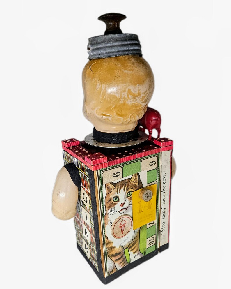 May include: A mixed-media sculpture featuring a vintage doll head topped with a dark wooden knob and a metal lid. The rectangular body is covered in game board graphics, including a cat illustration, the number 64, and the text "Moo, moo, says the cow."