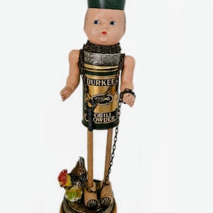 May include: A mixed-media doll figure featuring a vintage Durkee Chili Powder tin as the torso, a pale doll head with a green cap, thin wooden legs, and a small painted chicken figurine on a round wooden base, all connected by a metal chain.