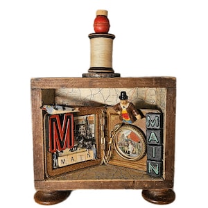 May include: A wooden shadow box display featuring a vintage spool topper, a map-lined interior, a miniature figure in a top hat, a circular framed image, and black letter tiles spelling MAIN. The display includes a red wooden finial on top.