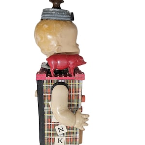 May include: A mixed-media assemblage sculpture featuring a doll head topped with a dark brown knob and a gray metal lid. A small red plastic pig sits on a plaid-covered rectangular base. A doll arm extends from the side, with letters N and K attached.