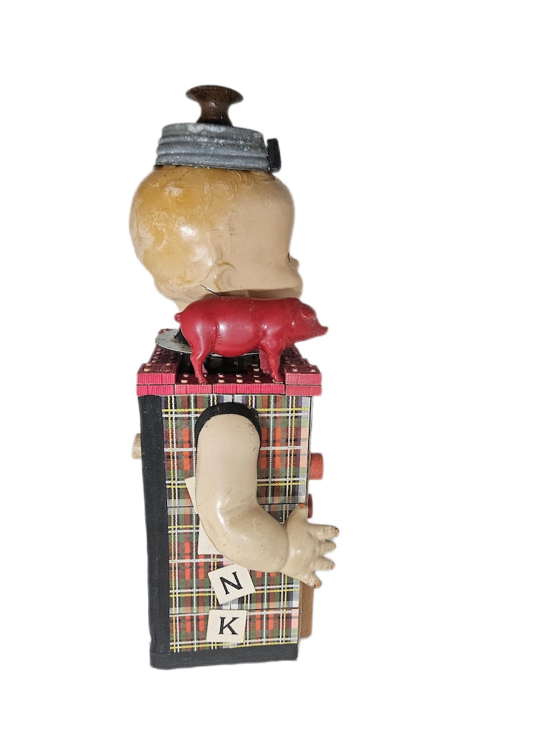May include: A mixed-media assemblage sculpture featuring a doll head topped with a dark brown knob and a gray metal lid. A small red plastic pig sits on a plaid-covered rectangular base. A doll arm extends from the side, with letters N and K attached.