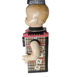 May include: A doll head mounted on a rectangular box base with a plaid pattern. The box features a protruding plastic arm and small square tiles with the letters O, I, N, K arranged vertically. The head is topped with a metallic lid and knob.