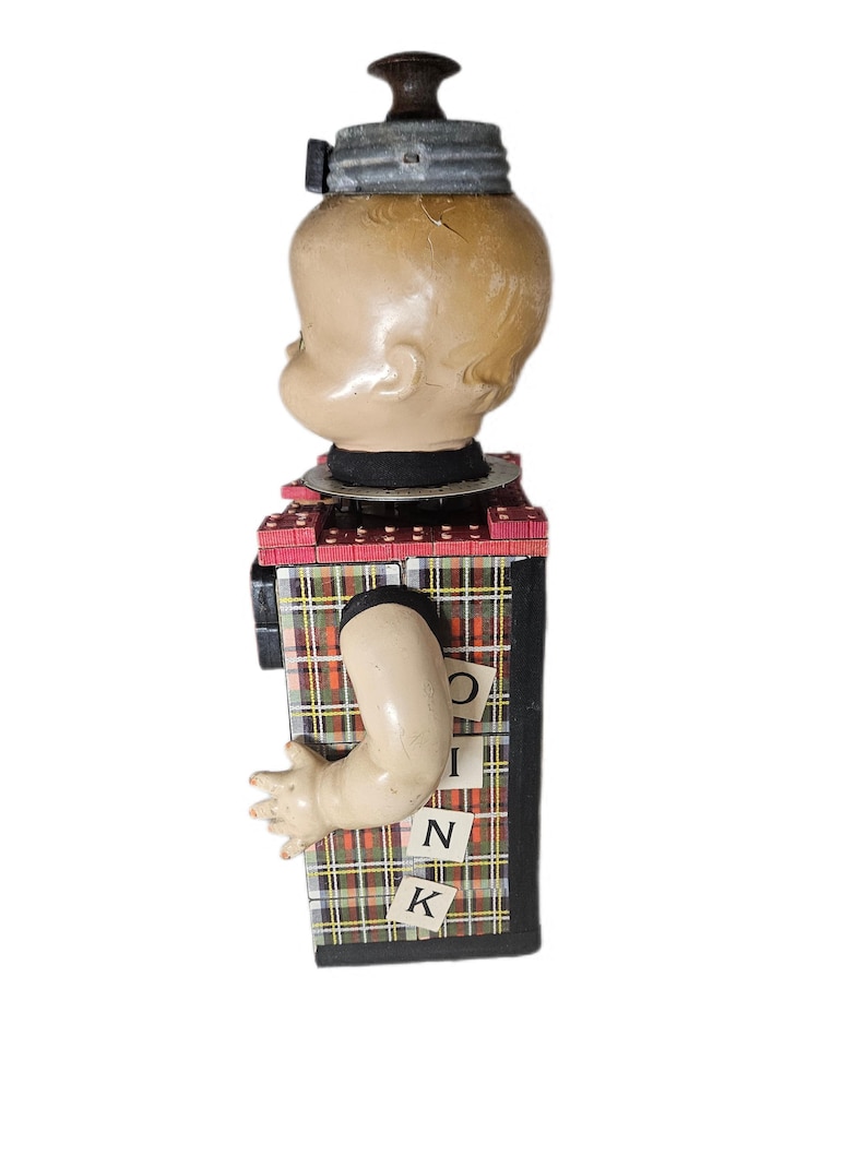 May include: A doll head mounted on a rectangular box base with a plaid pattern. The box features a protruding plastic arm and small square tiles with the letters O, I, N, K arranged vertically. The head is topped with a metallic lid and knob.