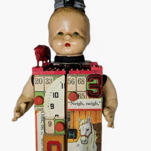 May include: A mixed-media doll sculpture featuring a vintage doll head topped with a gray cap and letter F, a red pig figurine, and a rectangular body constructed from blocks, bingo cards, a clock face, and a horse illustration with the text Neigh, neigh, says the horse.
