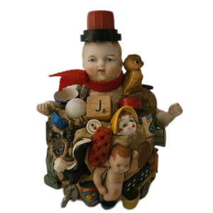 May include: A decorative assemblage sculpture featuring a central porcelain doll head wearing a red hat and scarf, surrounded by various miniature objects including a wooden J8 Scrabble tile, a small bird figurine, buttons, and doll parts.