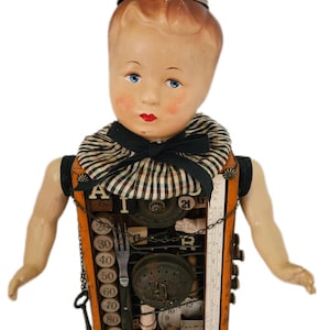 May include: A doll with a painted face and blue eyes, wearing a black and white striped ruffled collar. The torso is a rectangular wooden box filled with vintage objects including a metal fork, a ruler, numbered dials, and alphabet blocks.