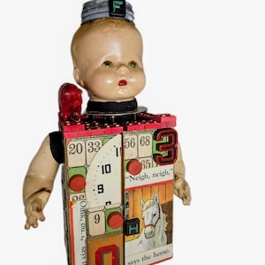 May include: A mixed-media doll sculpture featuring a vintage plastic doll head topped with a metal lid and the letter F. The rectangular body is constructed from wooden blocks decorated with bingo cards, a horse illustration, and the text "Neigh, neigh," says the horse and "Oink, ou," says the pig. It includes a small red pig figurine, the number 3, the number 0, and red wooden circles.