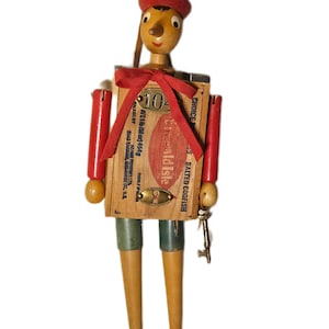 May include: A wooden figure with a red hat featuring a P3 tile, a long nose, and a torso made from an Emerald Isle Salted Codfish box. The figure has red arms, green shorts, and black shoes with 38 and 77 labels. A red ribbon is tied at the neck.