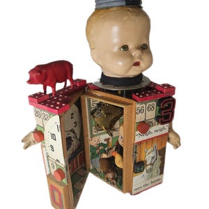 May include: A mixed-media sculpture featuring a doll head topped with a metal lid and the letter F. The body is a wooden box structure decorated with bingo cards, a red pig figurine, a clock face, and text including "Neigh, neigh," says the horse.