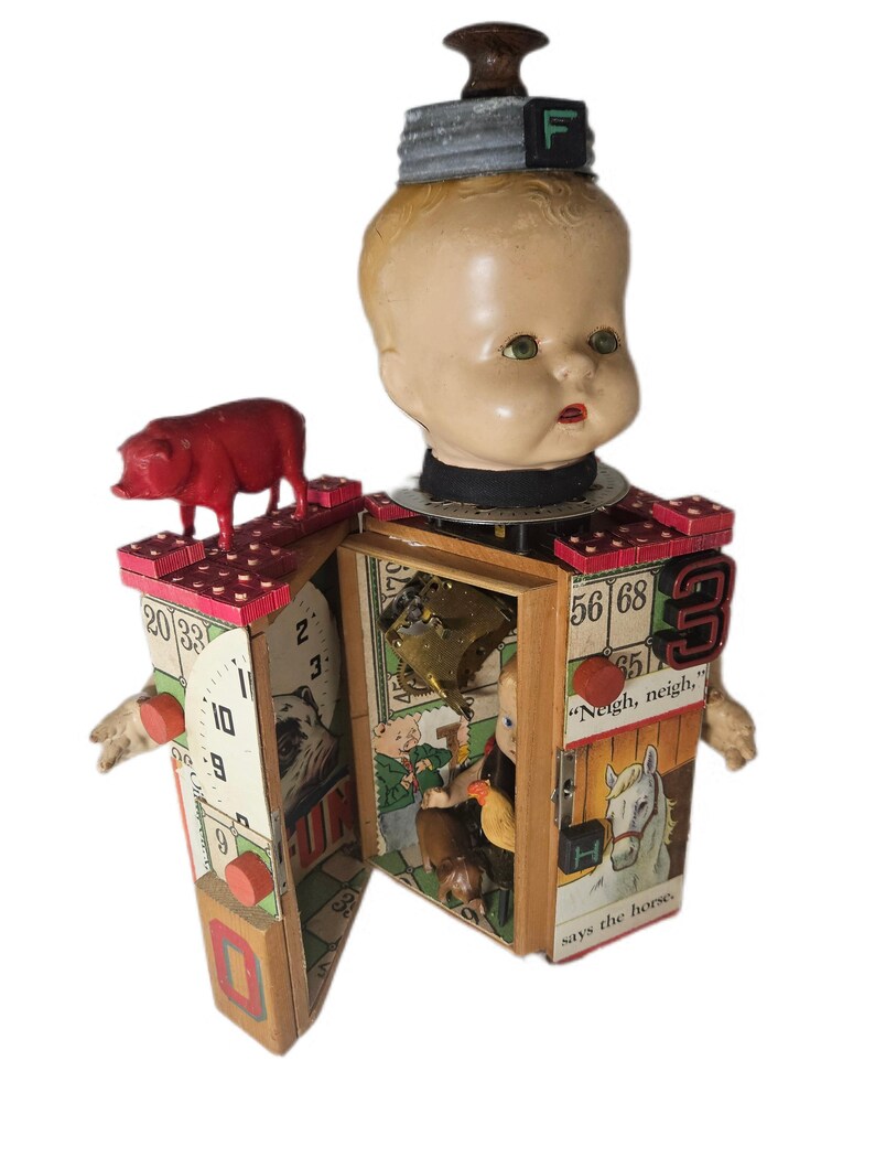 May include: A mixed-media sculpture featuring a doll head topped with a metal lid and the letter F. The body is a wooden box structure decorated with bingo cards, a red pig figurine, a clock face, and text including "Neigh, neigh," says the horse.
