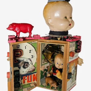 May include: A mixed-media assemblage sculpture featuring a large doll head atop a wooden box structure. The box is decorated with game board graphics, a red plastic pig, a small doll, a miniature chicken, and a brown pig figure inside.