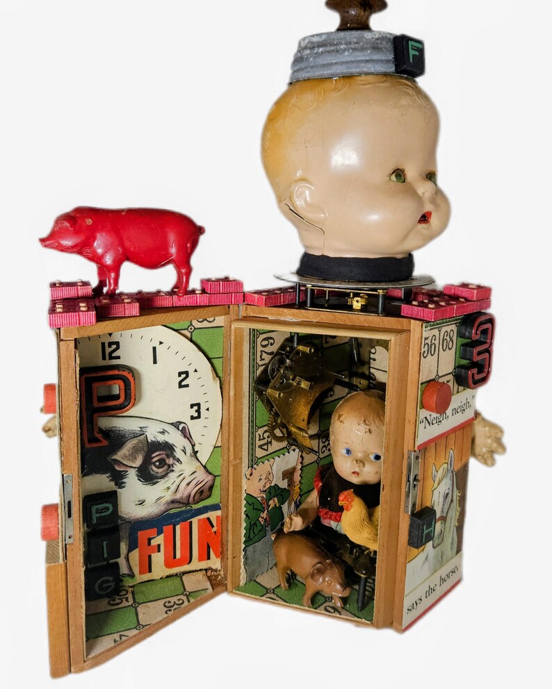 May include: A mixed-media assemblage sculpture featuring a large doll head atop a wooden box structure. The box is decorated with game board graphics, a red plastic pig, a small doll, a miniature chicken, and a brown pig figure inside.