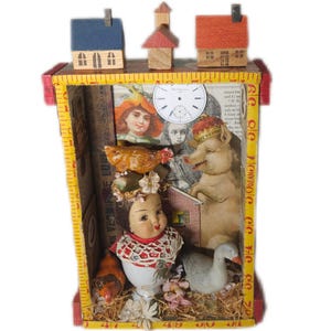May include: A whimsical shadow box with a yellow tape measure frame. Inside, a vintage doll, a goose, and a pig are surrounded by miniature houses and antique illustrations. The box is decorated with floral accents and a clock face.