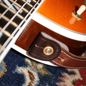 Puede incluir: Primer plano de una correa de guitarra de cuero marrón unida a una guitarra eléctrica de estilo vintage con una encuadernación blanca y herrajes dorados. La guitarra tiene un acabado sunburst y descansa sobre una alfombra estampada.