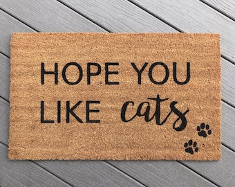 May include: A brown coir doormat with the text "HOPE YOU LIKE cats" in black lettering. There are three black paw prints in the bottom right corner.