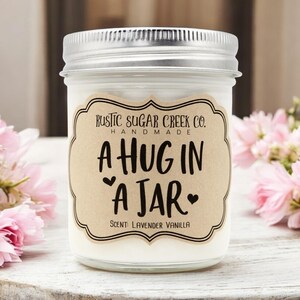 Op de afbeelding: Een handgemaakte kaars in een glazen pot met een zilveren deksel. Het etiket luidt "A HUG IN A JAR" met "Rustic Sugar Creek Co." en de geur "Lavender Vanilla". Roze bloemen staan op de achtergrond.