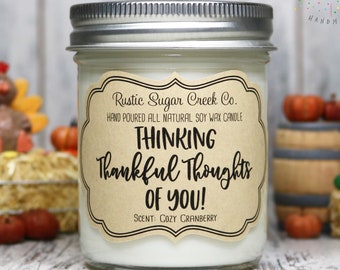 May include: A glass jar candle with a silver lid, labeled "Rustic Sugar Creek Co." and "Thinking Thankful Thoughts of You!" The candle has a cozy cranberry scent. Decorative pumpkins and a turkey figurine are in the background.
