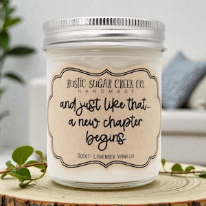 May include: A glass jar candle with a silver lid, resting on a wooden slice. The label reads "Rustic Sugar Creek Co. Handmade" and "and just like that... a new chapter begins." The scent is Lavender Vanilla.