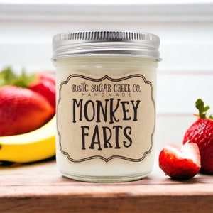 May include: A glass jar candle with a brown label that reads "Rustic Sugar Creek Co. Handmade Monkey Farts". The candle is sitting on a wooden surface with fruit and a banana.