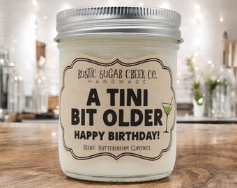 May include: A clear glass candle jar with a silver metal lid containing white wax. The label reads: RUSTIC SUGAR CREEK CO. HANDMADE A TINI BIT OLDER HAPPY BIRTHDAY! SCENT: BUTTERCREAM CUPCAKES, featuring a small green martini glass illustration.