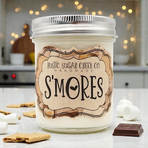 May include: A glass jar candle with a silver lid and a label that reads "Rustic Sugar Creek Co. Handmade S'mores." The candle is surrounded by graham crackers, marshmallows, and a piece of chocolate. The candle has a creamy white wax.