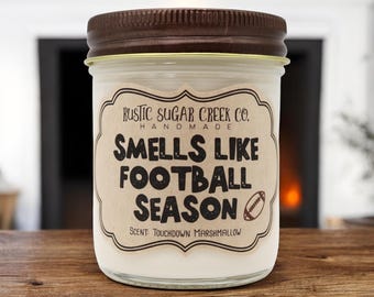 May include: A glass jar candle with a brown lid. The label reads "Smells Like Football Season" with a football graphic. The scent is "Touchdown Marshmallow". The candle is from Rustic Sugar Creek Co.