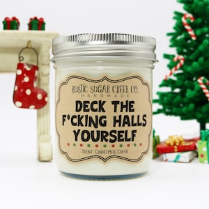 May include: A glass jar candle with a brown label that reads "RUSTIC SUGAR CREEK CO. HANDMADE DECK THE F*CKING HALLS YOURSELF" and "SCENT: CHRISTMAS CHEER".