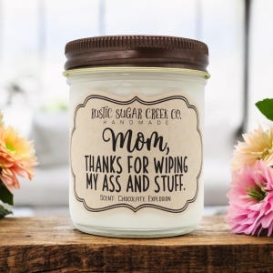 May include: A glass jar candle with a brown lid and a label that reads "RUSTIC SUGAR CREEK CO. HANDMADE Mom, THANKS FOR WIPING MY ASS AND STUFF. SCENT: CHOCOLATE EXPLOSION"